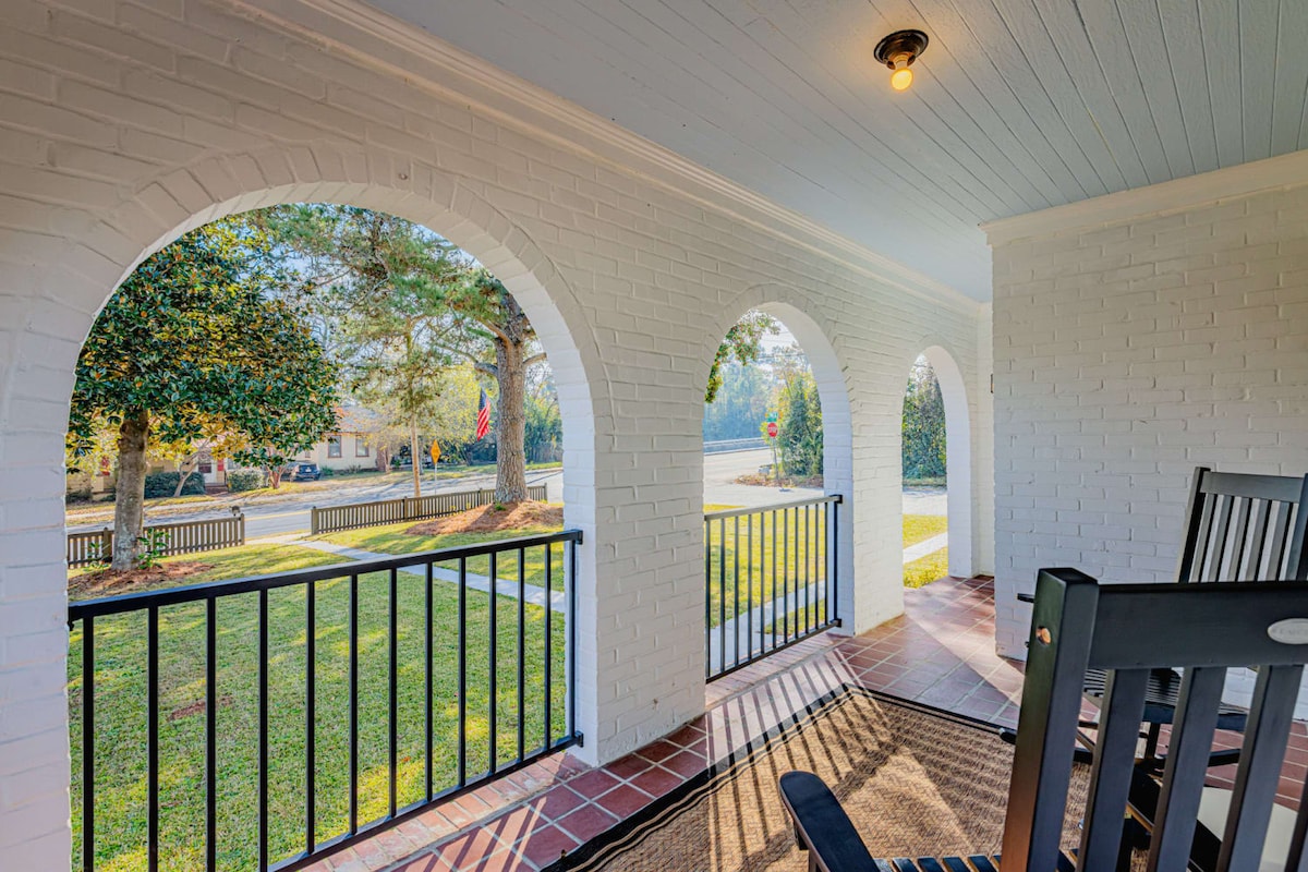 A welcoming porch is framed by three archways, revealing lush greenery and a tranquil neighborhood view. Rocking chairs provide a cozy spot to enjoy the fresh air, while the brick flooring adds classic charm. The soft morning light enhances the serene ambiance of this outdoor space.