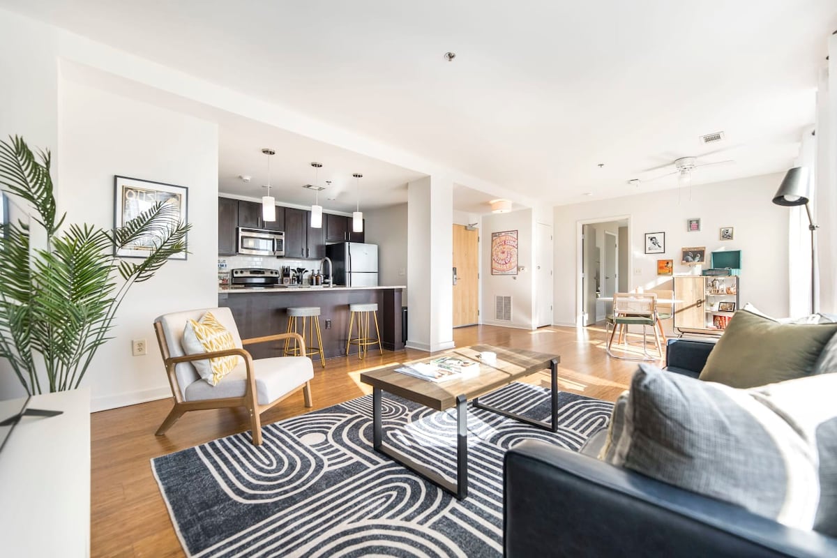 An open-concept living area features a modern kitchen with stainless steel appliances and a dining area with bar stools. A comfortable seating arrangement includes a dark couch and an accent chair on a patterned rug, illuminated by natural light from large windows.