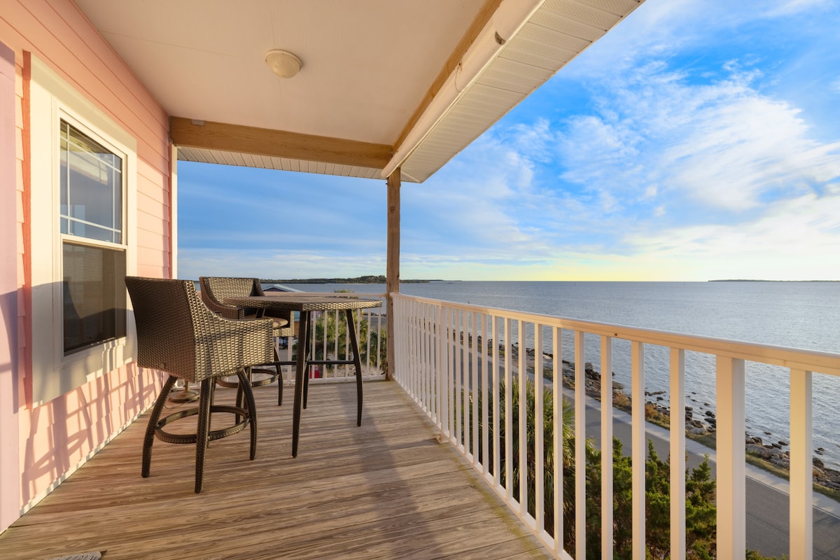 A covered deck extends from the upper unit, featuring outdoor seating with a dining table overlooking the Gulf. The expansive waterfront view captures the calm water and distant horizon, creating an inviting space for relaxation and sunset viewing.