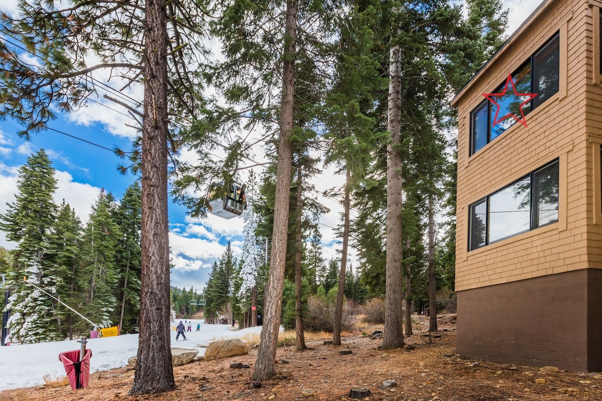 A snowy scene captures the exterior of a condo, surrounded by tall pine trees. The adjacent ski lift transports visitors, while skiers can be seen enjoying the slopes nearby. The building features large windows, allowing for scenic views of the winter landscape.