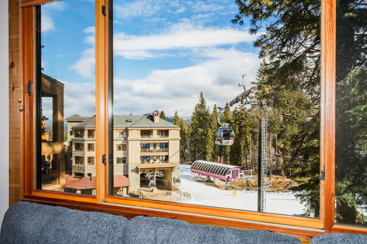 A large window frames a view of the snowy landscape, featuring the Big Springs Express Gondola and the Village Express lift. The scene includes snow-covered terrain and surrounding trees, complementing the structure of nearby buildings.