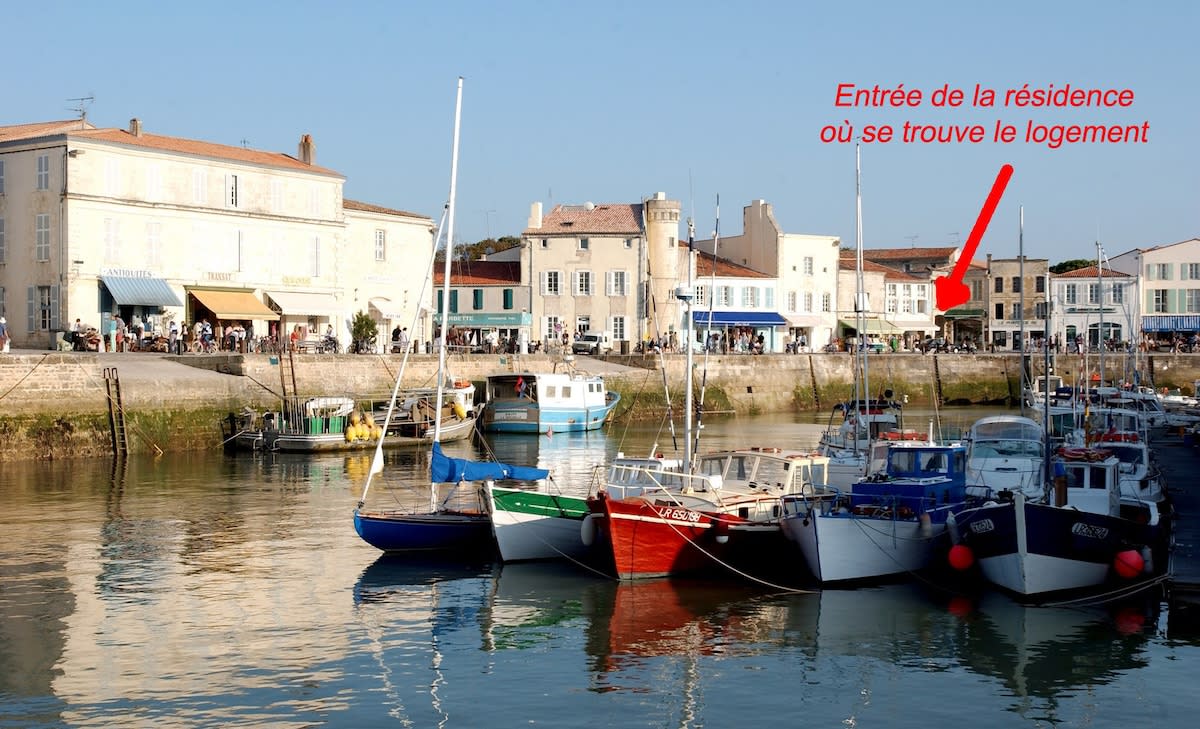 The image displays a picturesque marina scene with several boats docked in the foreground. In the background, charming buildings are visible along the waterfront, indicating the location of the residence. The serene water reflects the surrounding architecture, emphasizing the inviting coastal atmosphere.
