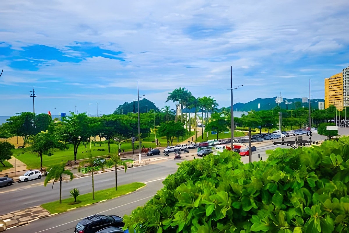 A street view is presented, showcasing a lively boulevard lined with shrubs and tall palm trees. The beach can be seen in the background, along with distant mountains under a partly cloudy sky. Various cars are parked along the road, indicating a vibrant atmosphere.