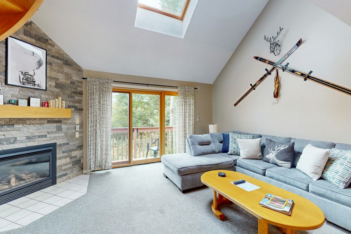 A sunlit living area features a high ceiling with a skylight, enhancing its spacious feel. A cozy gas fireplace is framed by a stone wall, while a sectional sofa sits across from a wood coffee table. Large glass doors lead to a balcony with views of greenery.