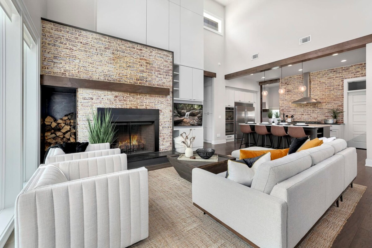 A spacious living area features a striking brick accent wall with a modern fireplace and a cozy seating arrangement. Two light-colored sofas and a textured area rug enhance the space. The open layout connects to a stylish kitchen with bar seating and high-end appliances.