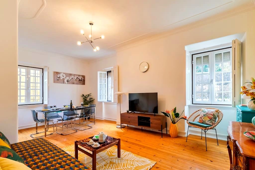 A bright living space is presented featuring a television and a dining area with a glass table and chairs. Windows allow natural light to fill the room, highlighting the wooden floor. Decorative elements such as a patterned sofa and potted plants add visual interest.