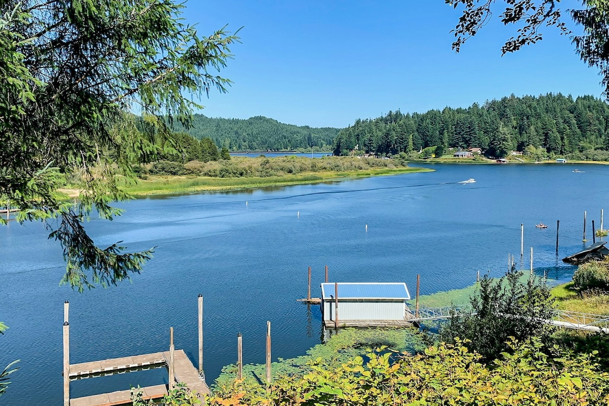 A serene lake view is displayed, with calm waters reflecting the clear blue sky. A private boathouse and wooden dock are visible in the foreground, surrounded by lush greenery and gently sloping hills, creating a peaceful atmosphere. Distant trees line the opposite shore.