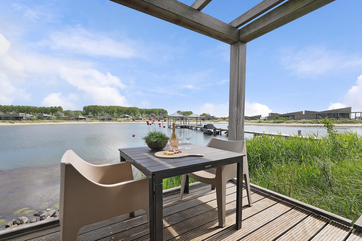 An outdoor dining area features a table set with glassware and a small plant. Two chairs are positioned around the table, with a view of the water and surrounding greenery in the background, enhancing the serene atmosphere.