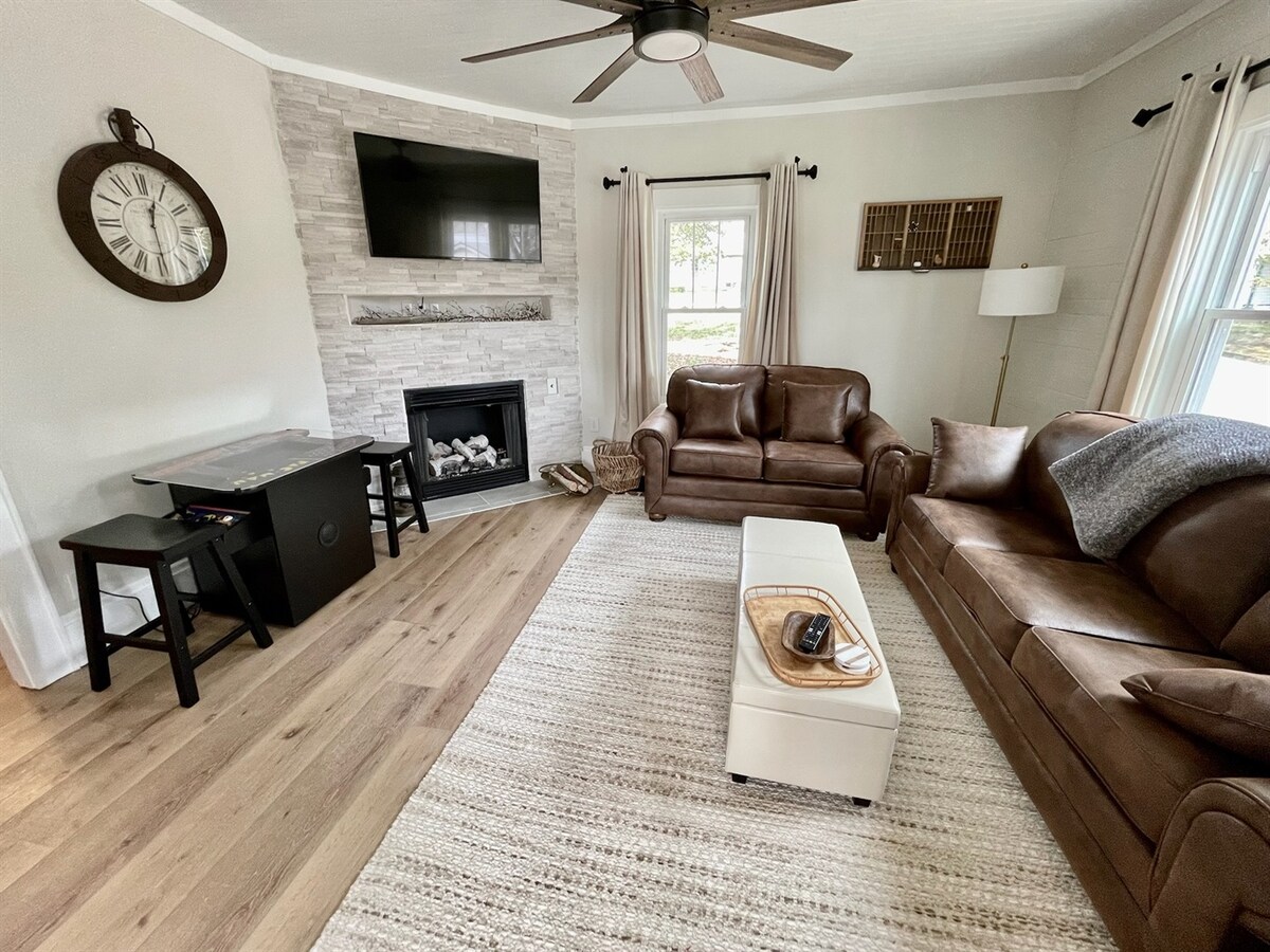 A living room features two leather sofas facing a modern fireplace, complete with a TV mounted above. A wooden coffee table sits in the center, while a small black table is positioned beside. Light streams in from a window dressed with curtains, adding warmth to the space.