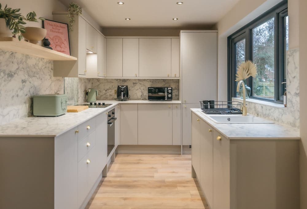 A modern kitchen features light cabinetry and marble countertops. Natural light filters through large windows, illuminating the space. Equipped with essential appliances, including an oven and coffee machine, the room promotes a functional cooking environment with ample counter space for meal preparation.