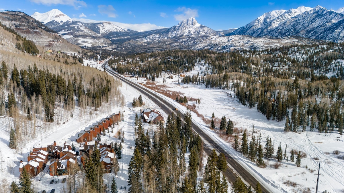 A scenic aerial view captures a collection of rustic condos nestled among a snowy landscape. Tall evergreen trees and snow-covered mountains frame the horizon, with a winding road leading through the serene winter scenery.