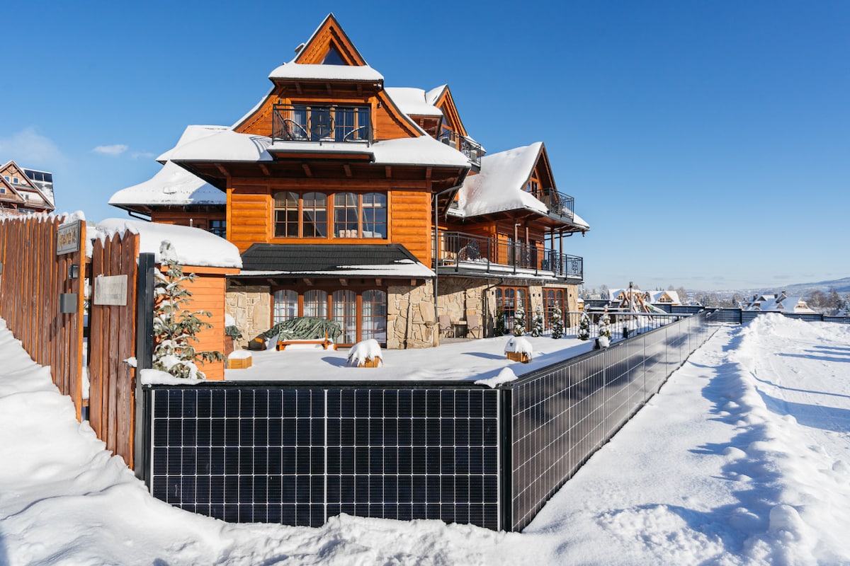 The architectural design features a charming wooden structure set against a clear blue sky, surrounded by a snowy landscape. Solar panels are visible on the building's lower level, and a fenced area complements the serene environment.