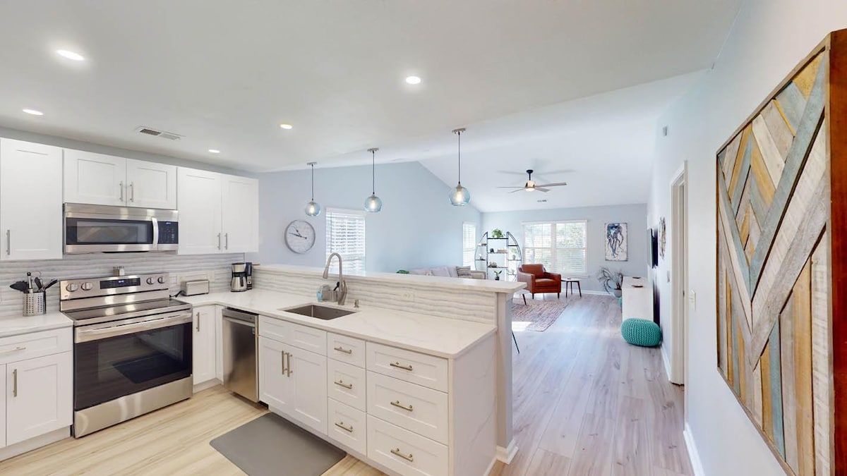 A modern kitchen features stainless steel appliances and a spacious island with seating. Light-colored cabinets complement the sleek countertops. In the background, a bright living area is visible with ample natural light streaming through large windows.