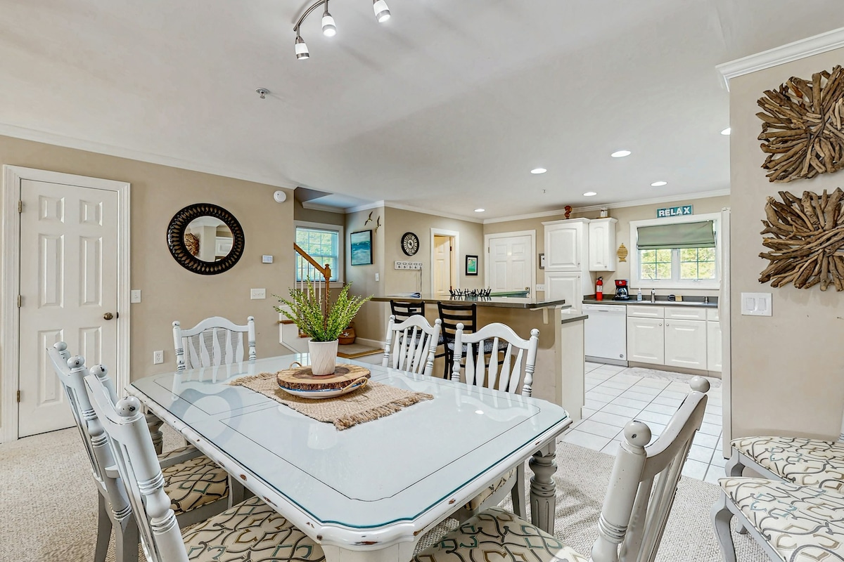 A bright dining area features a large white table surrounded by six chairs, set on a patterned rug. In the background, a well-lit kitchen with white cabinetry and modern appliances is visible, creating an open and inviting space.