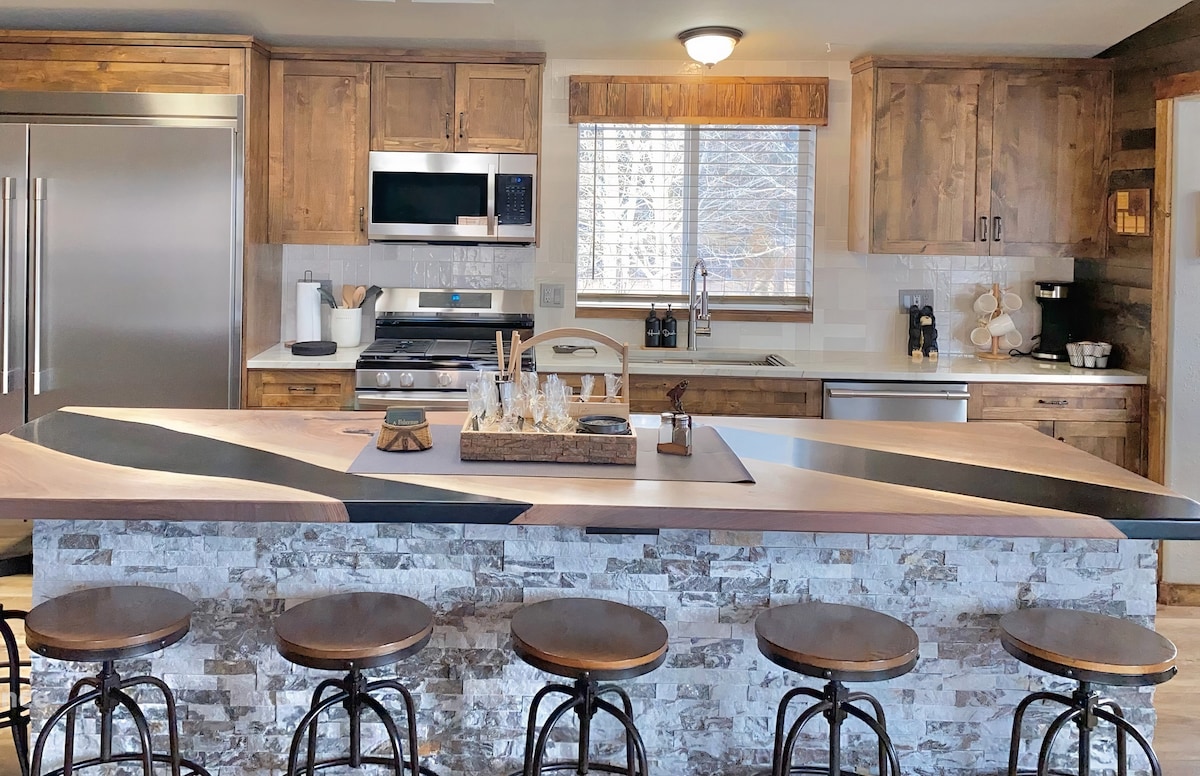 A modern kitchen features wood cabinetry and stone accents. Stainless steel appliances are visible alongside a double refrigerator. A large island with a wooden countertop is surrounded by six metal stools, providing ample seating for dining or socializing.