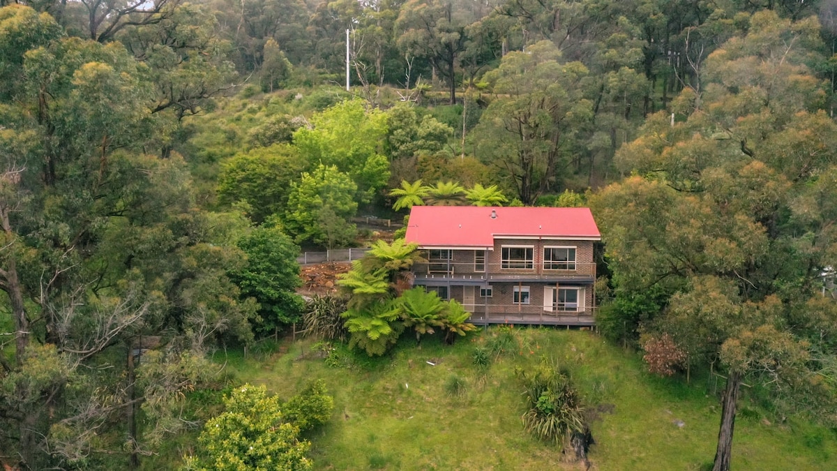 The house is nestled among lush greenery, featuring a red roof that contrasts with the surrounding trees. Large windows and a spacious deck are visible, blending the residence harmoniously with the natural landscape.