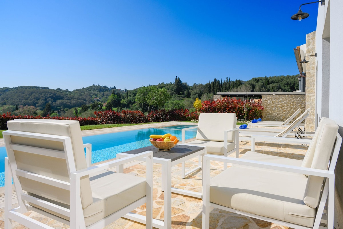 An outdoor seating area features two white chairs positioned beside a stone patio, overlooking a sparkling pool. A bowl of fresh fruit rests on a small table. Lush greenery and colorful foliage create a serene backdrop against the clear blue sky.