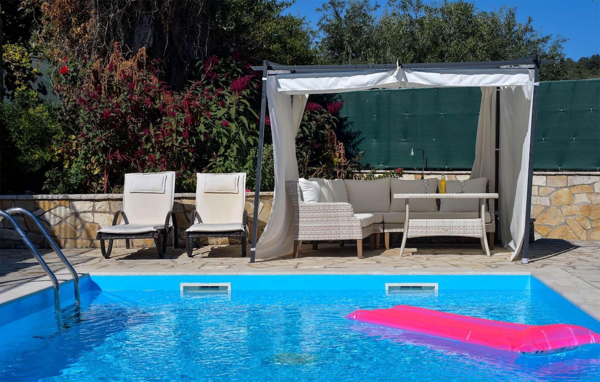 A private pool is surrounded by a stone patio, featuring a seating area with a canopy and a table. Two loungers are positioned nearby, and vibrant greenery provides a serene backdrop. A floating inflatable adds a splash of color to the water.