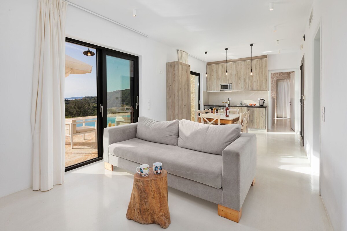 A bright living area features a light grey sofa accompanied by a rustic wooden side table. Large glass doors open to a terrace with views, allowing natural light to fill the space. A modern kitchen is visible in the background, with minimalistic cabinetry and pendant lighting.