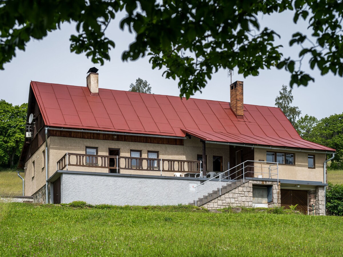 The semi-detached house features a red roof and a mix of stone and stucco exterior. A spacious balcony with a railing extends across the front, offering access to views of the surrounding greenery. Large windows allow for natural light, enhancing the countryside charm.