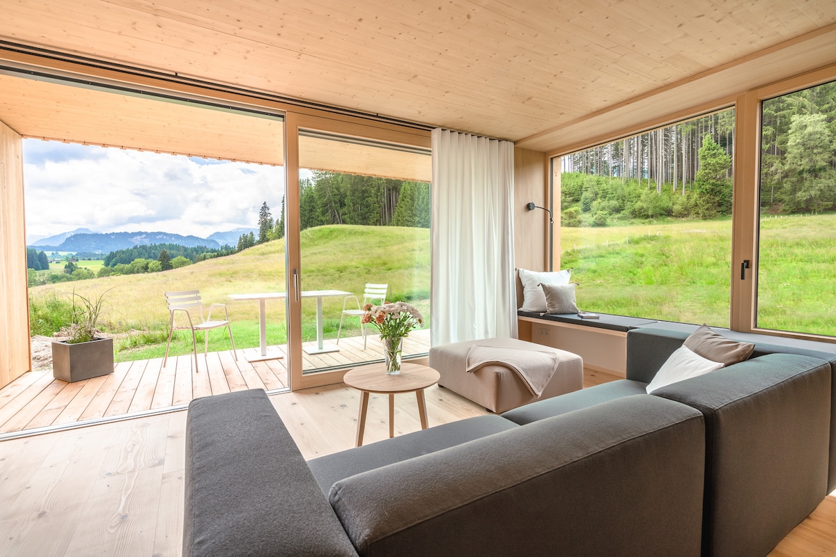 A bright living area features large glass doors opening to a patio with views of the surrounding greenery and mountains. A comfortable gray sofa faces a small round table, while a potted plant adds a touch of nature indoors. Natural light fills the space.