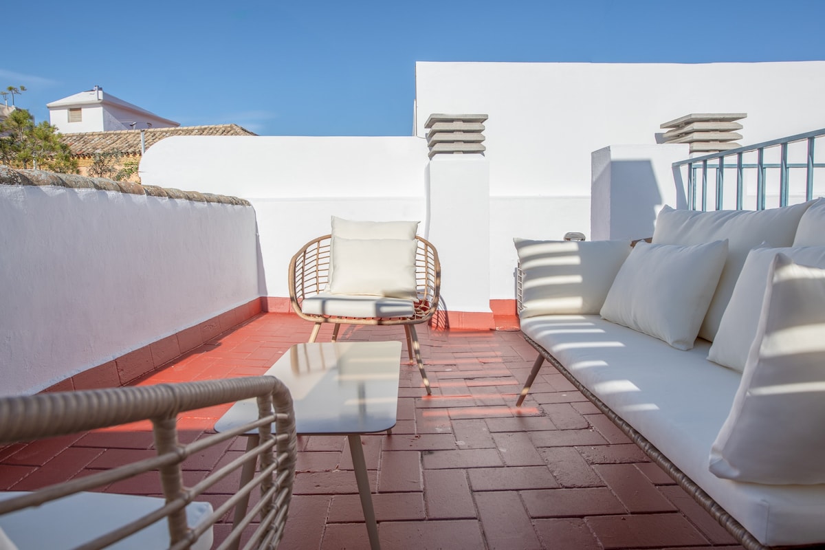 A private terrace is adorned with a comfortable seating area featuring two rattan chairs and a cushioned sofa. A small, modern table stands between them on the terracotta-tiled surface, providing a relaxing space to enjoy the sunshine and views.