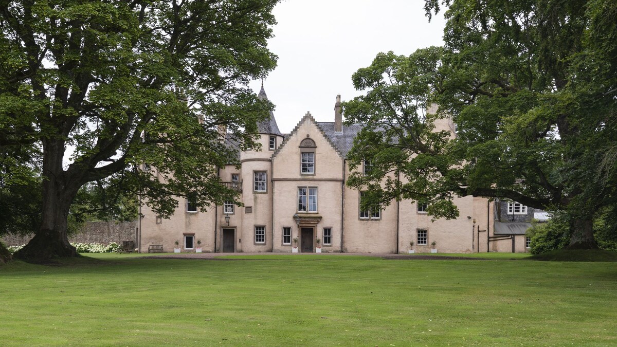 The stately facade of Keith Marischal House showcases its historic Scots Baronial architecture, framed by lush greenery and mature trees. A well-maintained grassy area extends in front of the house, creating an inviting outdoor space.