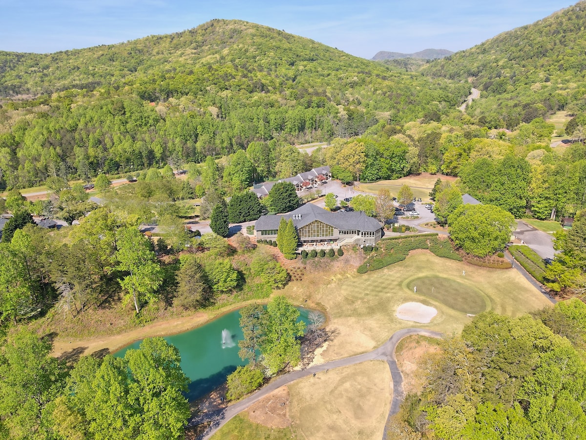 Aerial view of a lush landscape featuring a small pond with a fountain surrounded by greenery. A golf course can be seen nearby, alongside a cluster of buildings designed in harmony with the natural surroundings, set against the backdrop of rolling hills.
