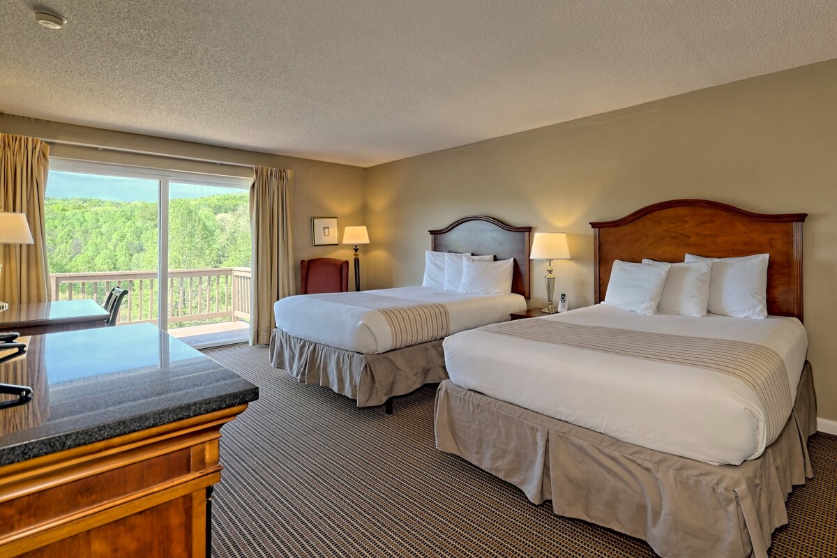 A spacious guest room features two Queen beds with soft linens and decorative headboards. Large glass doors lead to a private balcony with views of the surrounding greenery. Wood furnishings and lamps provide functionality, while neutral tones contribute to a calming environment.