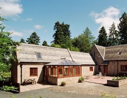 The exterior of the stone cottage is showcased, featuring a traditional slate roof and large windows. A glass conservatory connects the main building to a private patio area, surrounded by greenery and highlighted by blue skies and scattered clouds.