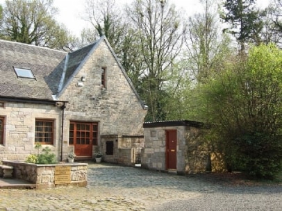 The exterior of a stone cottage is visible, showcasing a charming design with a steep roof. A cobbled courtyard leads to the entrance, framed by greenery from surrounding trees and shrubs. A small structure can be seen nearby, contributing to the tranquil setting.