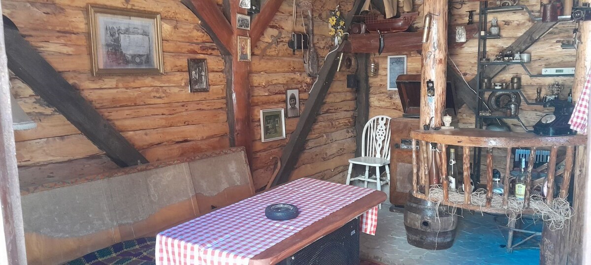 A rustic interior showcases wooden beams and paneling, creating a warm atmosphere. A checkered tablecloth covers a wooden table with a central decorative feature. Various vintage items and photographs are displayed on shelves, enhancing the room's character. A white chair complements the earthy tones.