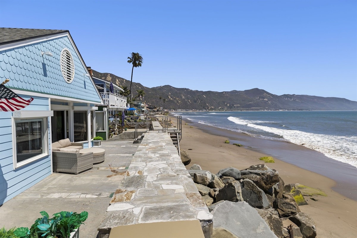 A coastal pathway extends alongside the beach, bordered by smooth stones. An inviting home with light blue siding is visible, featuring a circular window. The scene captures the sandy shore and rolling ocean waves under clear skies, with distant mountains lining the coastline.
