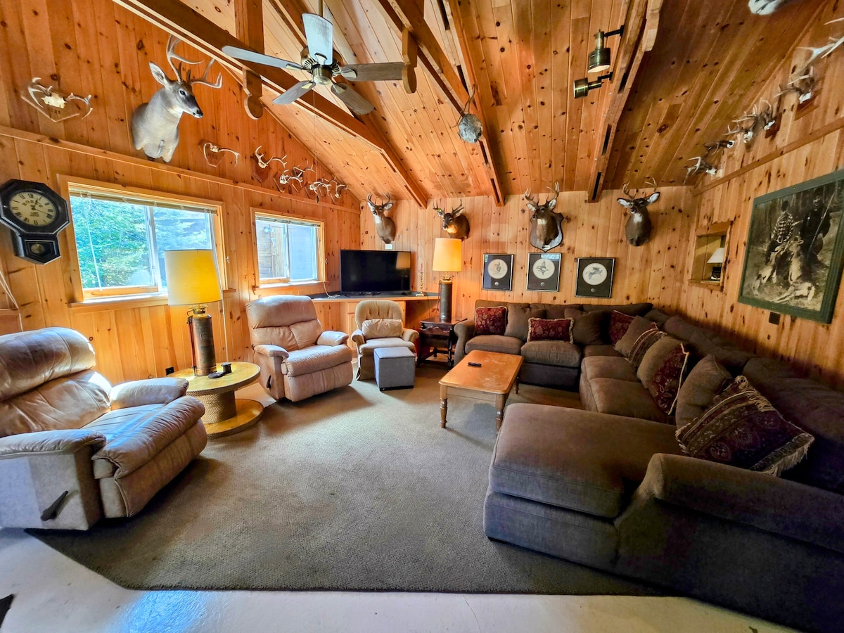 A spacious den features wood paneling and a vaulted ceiling, furnished with a large sectional sofa and several recliners. Deer antlers adorn the walls, while two windows illuminate the room. A flat-screen TV and a coffee table complete the inviting lounge area.