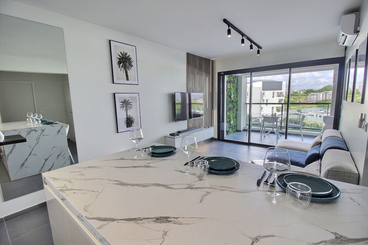 A modern living area features a sleek marble kitchen island with paired dinnerware set. Contemporary artwork adorns the walls, and large glass doors open to a terrace overlooking the marina, allowing natural light to fill the space.