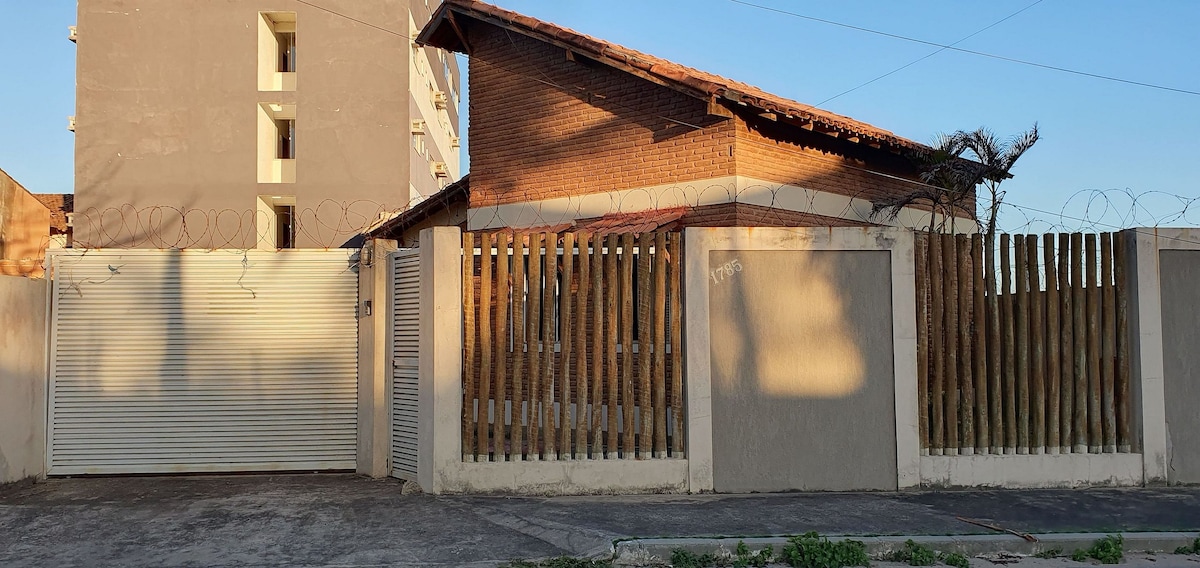 The exterior of the house is displayed, featuring a rustic brick facade and a sloped roof. A gate is visible, along with a fence made of wooden slats and concrete walls. A tall building stands in the background, indicating proximity to the surrounding area.