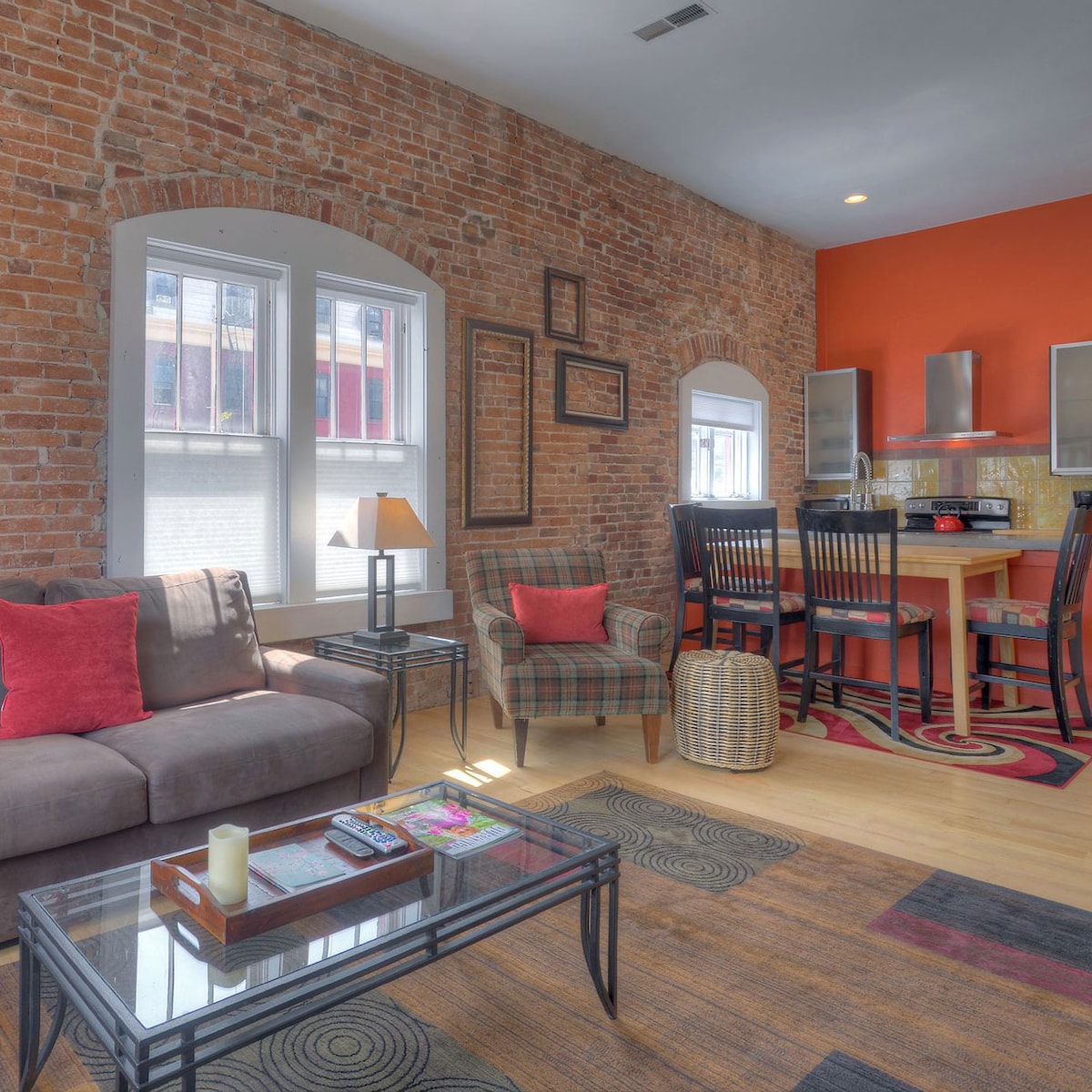 An open living space showcases exposed brick walls, accented by a vibrant orange feature wall. A comfortable sofa with red cushions is paired with a plaid armchair. A dining table is visible nearby, surrounded by black chairs, with modern appliances in the kitchen area.