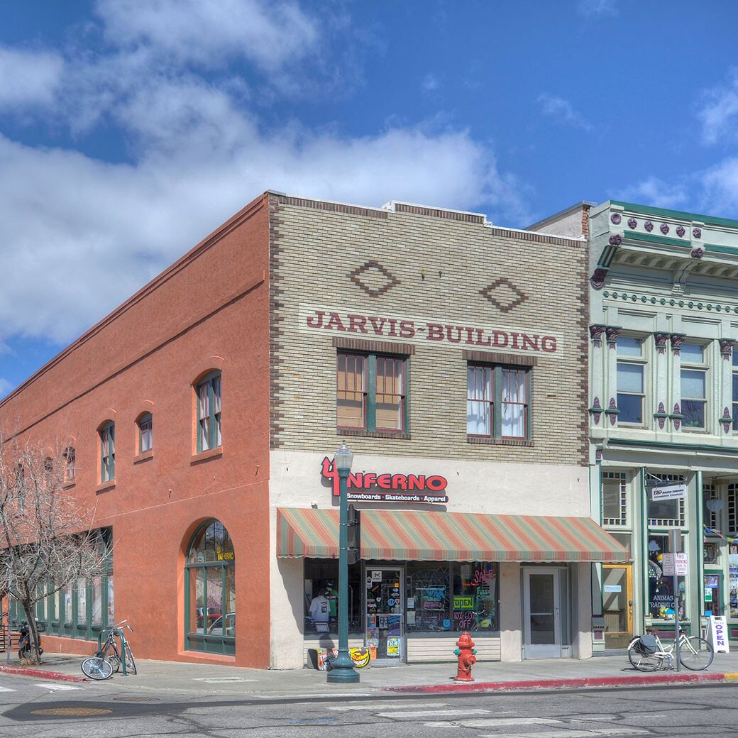 The exterior of the Jarvis Building is shown, featuring a mix of brick and painted facades. Large windows provide a view into the street-level businesses below. A striped awning extends over the entrance, while neighboring buildings exhibit distinct architectural details from different eras.