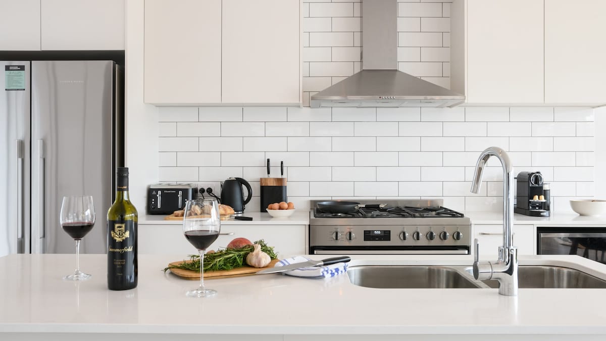 A modern kitchen features a spacious island counter with a bottle of wine and two glasses. Stainless steel appliances include a gas stove and refrigerator, complemented by a stylish backsplash. Fresh herbs and vegetables are displayed on a wooden cutting board, enhancing the inviting atmosphere.