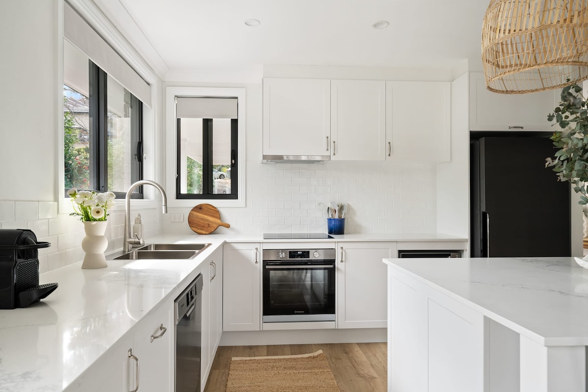 A modern kitchen featuring white cabinetry and a marble countertop. Stainless steel appliances, including an oven and dishwasher, are visible. A large window allows natural light, while a spacious sink is complemented by a stylish faucet. A woven pendant light adds a touch of warmth.
