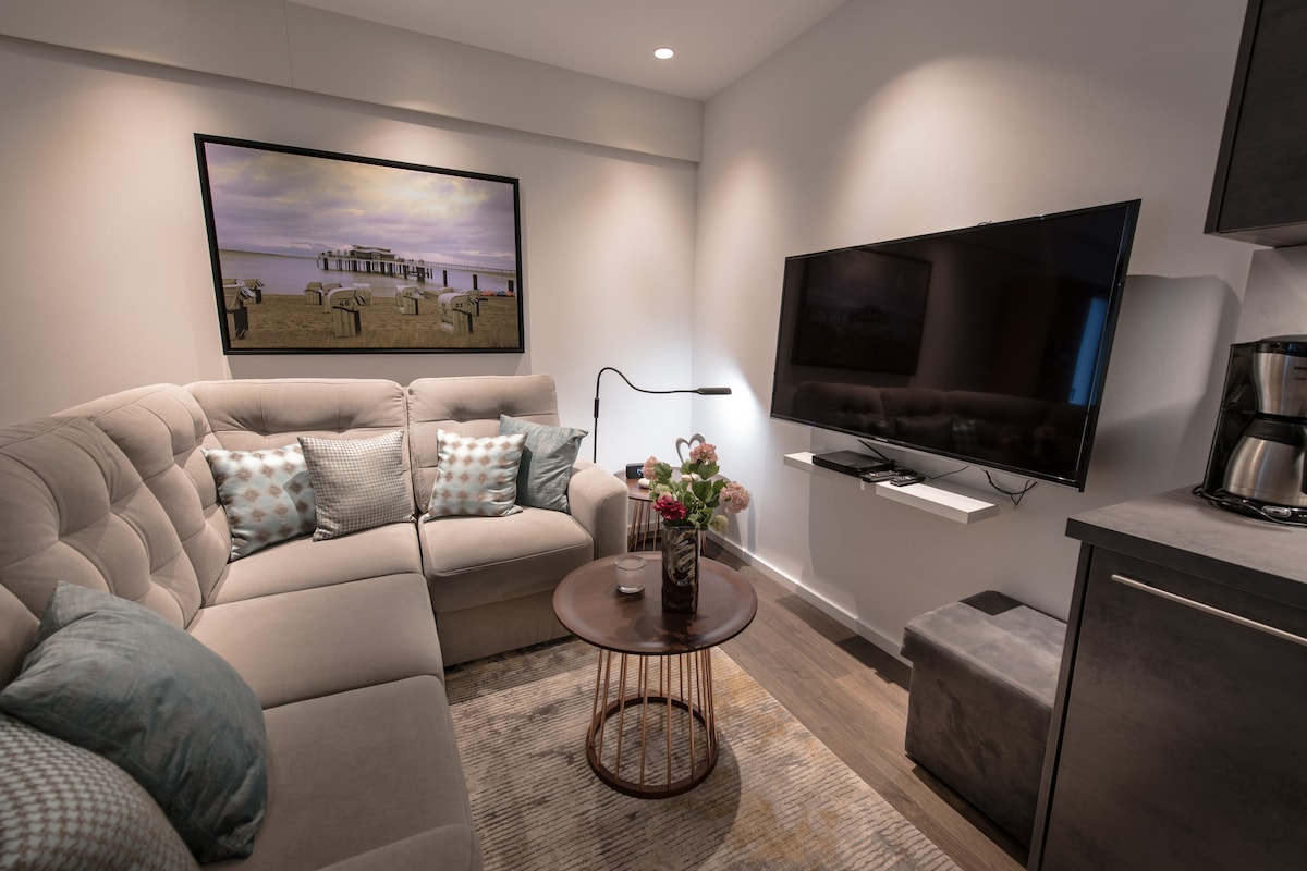 A modern seating area features a light-colored sectional sofa adorned with decorative pillows. A round table sits in front, complemented by a small vase of flowers. A flat-screen television is mounted on the wall, alongside a coffee maker integrated into the cabinetry.