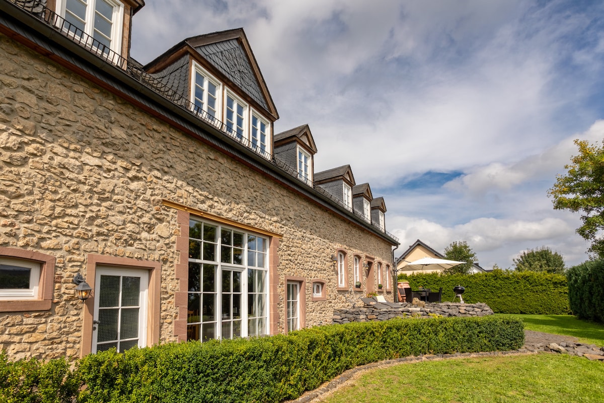 The exterior of a spacious stone building is featured, displaying multiple windows and a well-maintained garden area. A lush green landscape surrounds the property, and a stone pathway leads to the entrance. The structure is complemented by a classic architectural style.