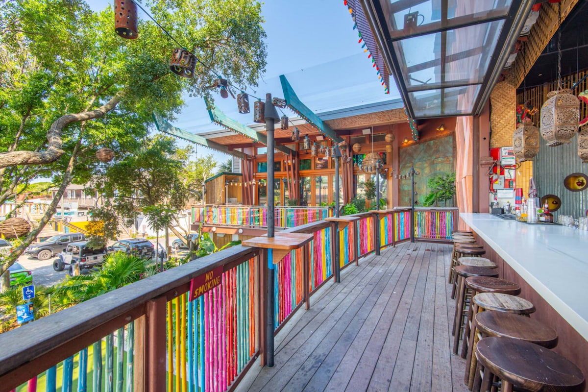 A vibrant outdoor bar area features a wooden deck with colorful railing accentuated by tropical decor. Stools are positioned along the bar, providing ample seating for guests. Lush greenery is visible in the background, contributing to a lively yet relaxed atmosphere.