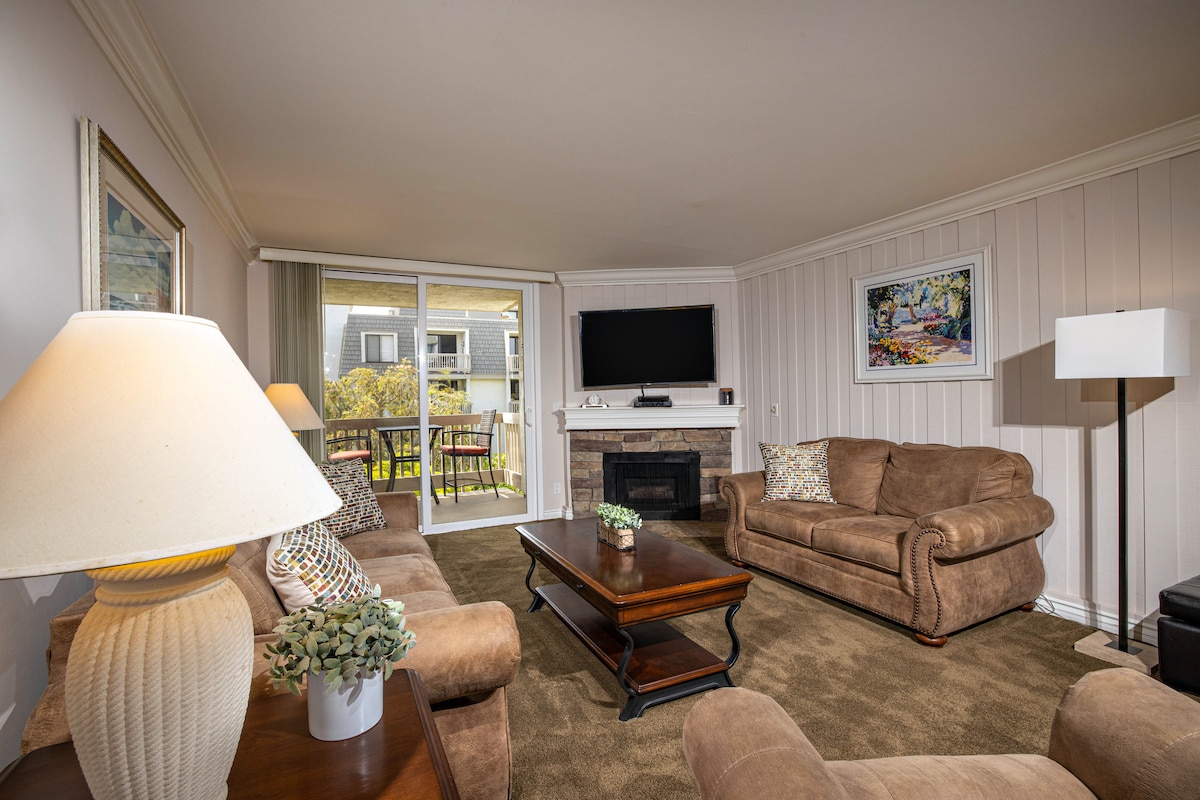 A spacious living area features comfortable seating arranged around a wooden coffee table. A television is mounted on the wall above a stone fireplace. Natural light streams through large sliding glass doors that lead to a balcony, overlooking landscaped grounds.
