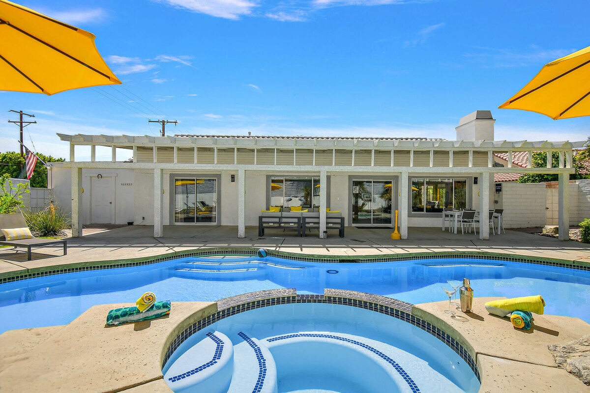 A spacious outdoor area surrounds a refreshing pool and spa. Shaded by yellow umbrellas, the poolside features a seating area with benches and a dining table. The sliding glass doors in the background provide access to the indoor living space and showcase a clear blue sky above.