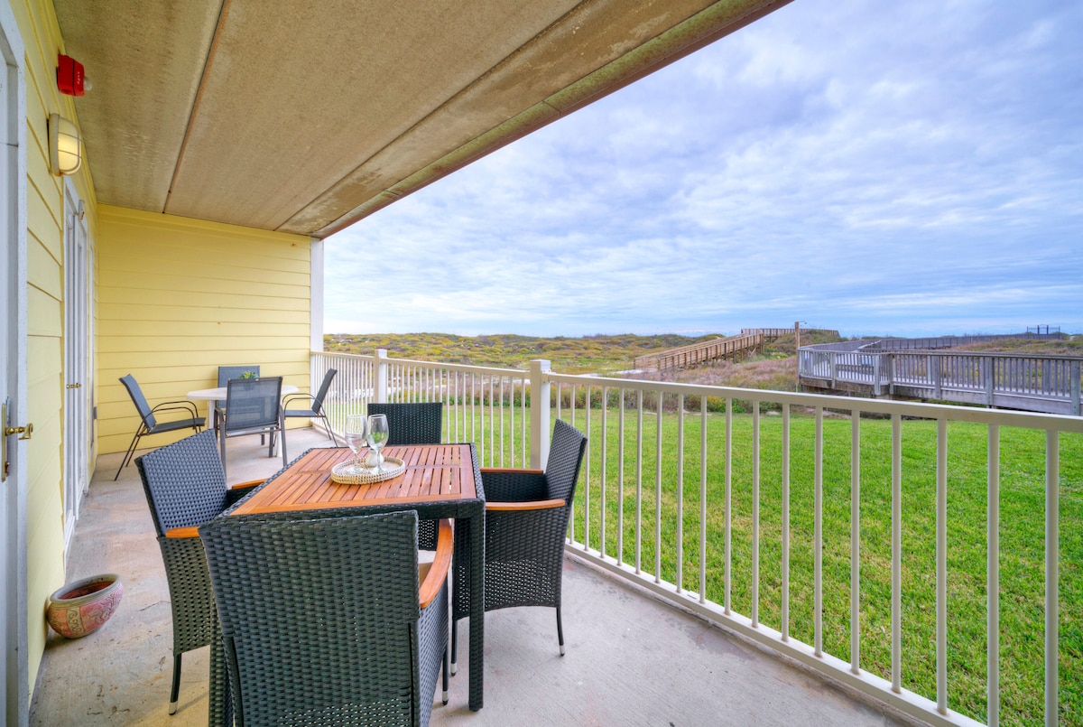 A covered private patio is shown, featuring dual al fresco dining tables surrounded by black wicker chairs. The patio offers unobstructed views of the lush green lawn and the boardwalk leading to the beach in the distance under a cloudy sky.