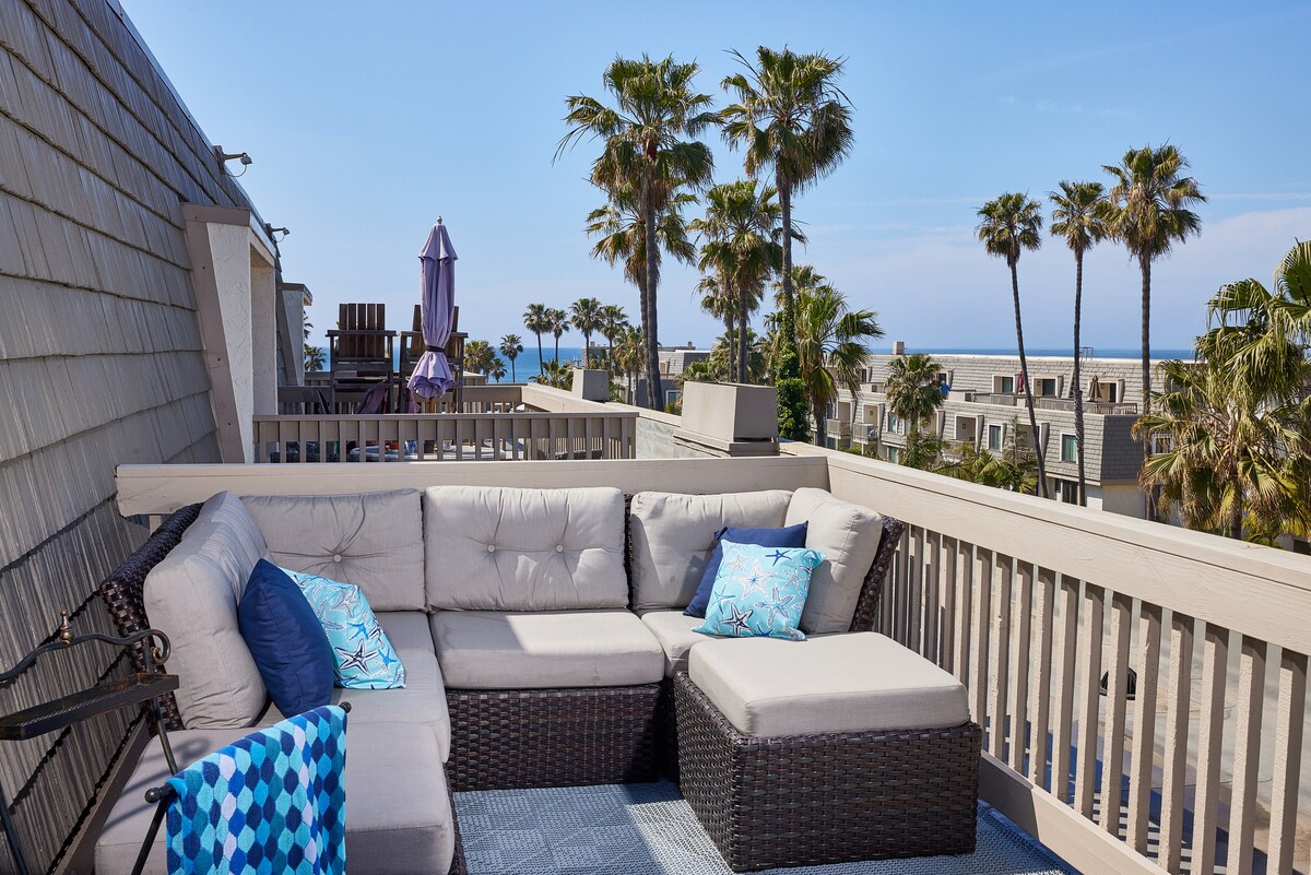 A spacious rooftop balcony features a large sectional sofa with light cushions and decorative pillows. Surrounding tropical palm trees and an ocean view create a serene outdoor space. A small table complements the seating arrangement, enhancing the inviting atmosphere.
