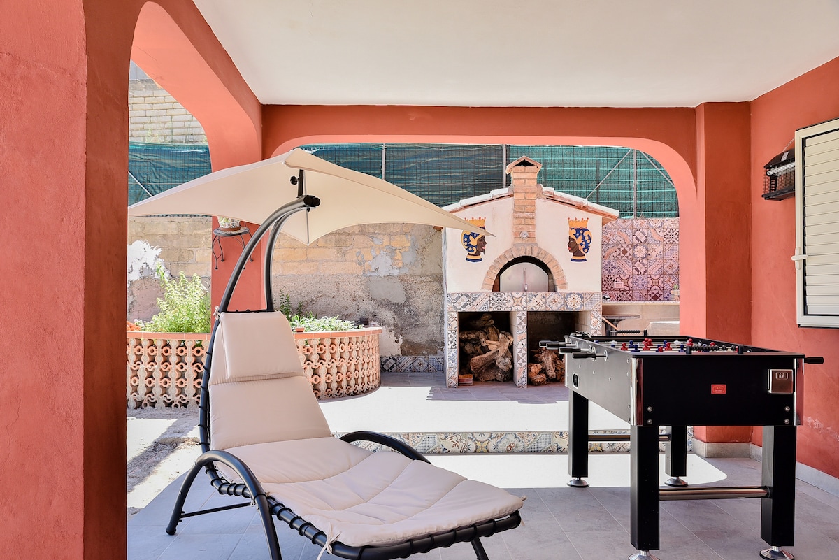 A shaded outdoor area features a lounge chair and a foosball table. A brick barbecue with decorative tiles is visible in the background, while vibrant red walls frame the space, enhancing its warmth and invitation for leisure activities.