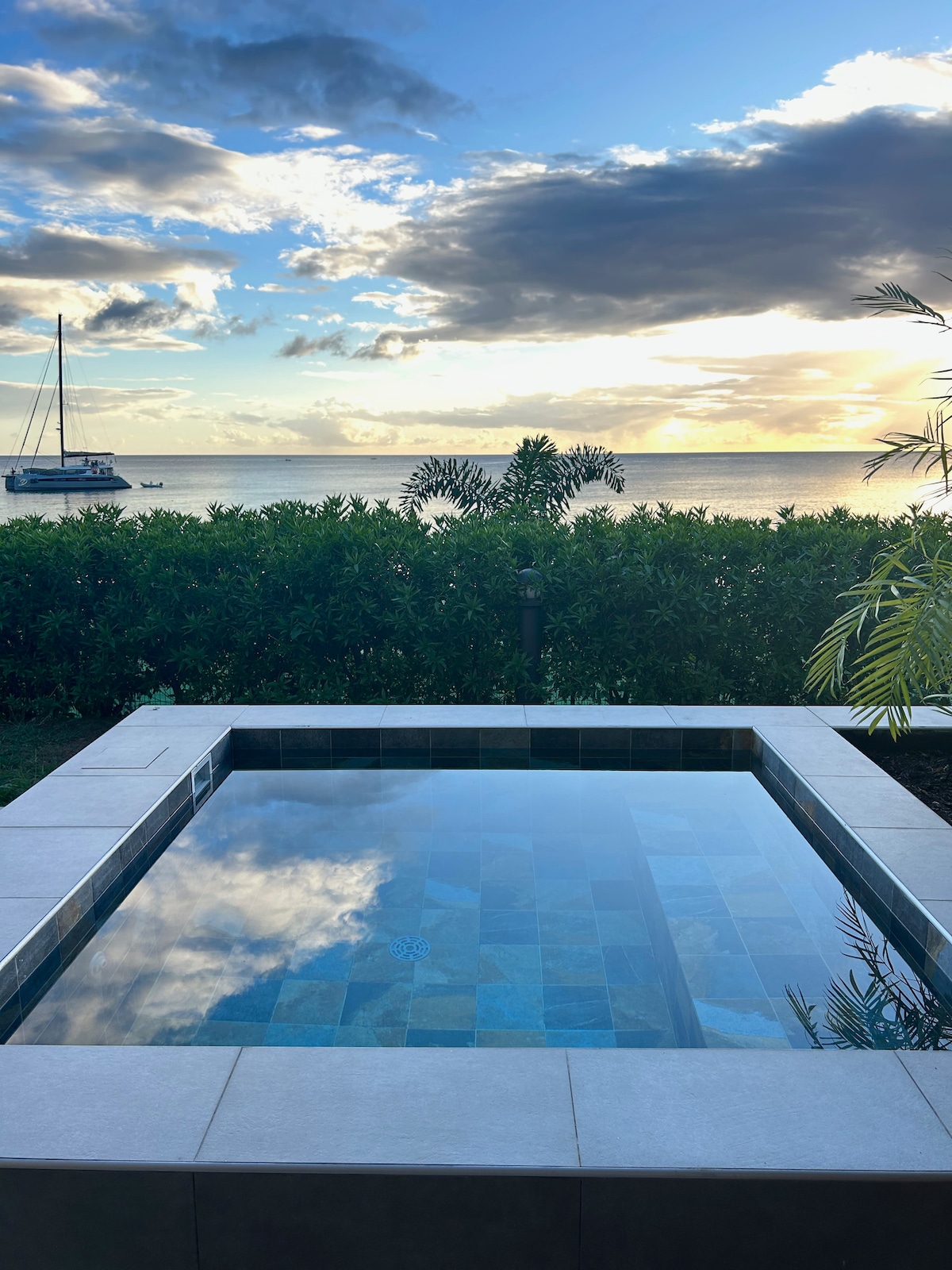 A serene plunge pool is framed by a paved area, reflecting the sky's hues above. Lush greenery borders the pool, with a view of the calm sea and a sailboat in the distance, enhancing the tranquil atmosphere of the space.