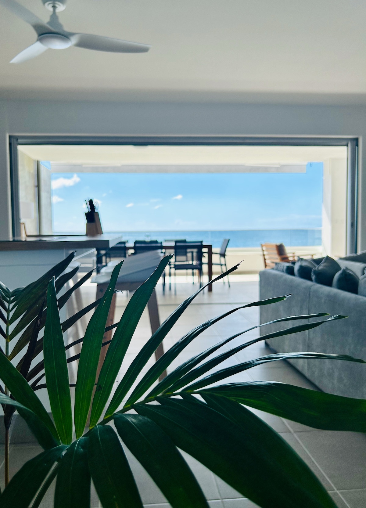 A spacious living area is visible, featuring large glass doors that open to a sea view. A dining table for six is positioned in the background, with a beachfront setting. Greenery from a nearby plant adds a touch of nature to the interior.